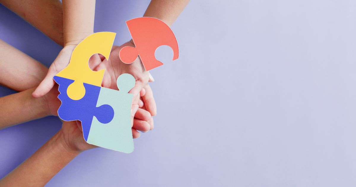Hands holding colorful puzzle pieces shaped like a human head on a purple background.